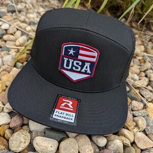 May include: Black flat bill snapback hat with a mesh back. The hat features a USA patch with a red, white, and blue design. The hat has a Richardson tag.