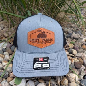 May include: A gray and black trucker hat with a leather patch that reads "SMITH FARMS EST.1970" with a tractor graphic. The hat has a curved brim and an adjustable strap.