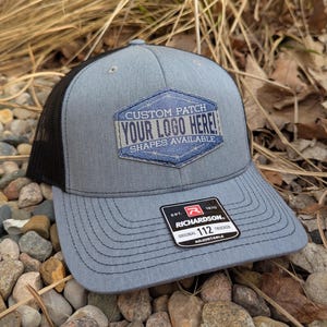 May include: A gray and black trucker hat with a custom patch that says "CUSTOM PATCH YOUR LOGO HERE! SHAPES AVAILABLE". The hat is made by Richardson and is adjustable.