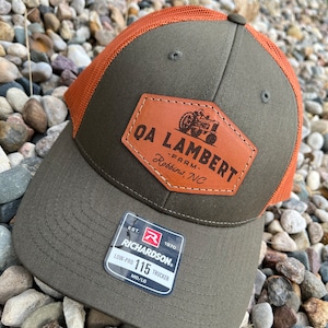 May include: A green and orange trucker hat with a brown leather patch that says "OA Lambert Farm, Robbins, NC". The patch features a tractor graphic. The hat has a Richardson 115 label.