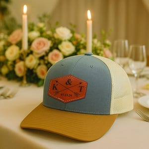 May include: A blue and beige baseball cap with a brown leather patch that says "K & T 05.13.24" in black lettering. The patch has two arrows crossing each other.