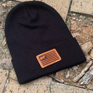 May include: Black knit beanie with a brown leather patch featuring a silhouette of a cow and a stylized American flag.