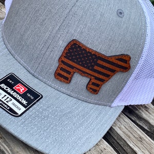 May include: A gray and white trucker hat with a brown leather patch featuring an American flag design. The patch is shaped like a cow. The hat has a Richardson logo and the text "EST. 1970" and "1 1/2 TRUCKER ADJUSTABLE".