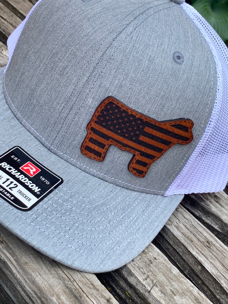 May include: A gray and white trucker hat with a brown leather patch featuring an American flag design. The patch is shaped like a cow. The hat has a Richardson logo and the text "EST. 1970" and "1 1/2 TRUCKER ADJUSTABLE".
