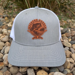 May include: A gray and white trucker hat with a curved brim. The hat features a brown leather patch with the words "WEEKEND HOOKER" and a graphic of a fish jumping out of the water, with crossed fishing rods.