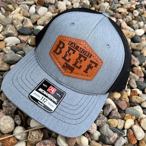 May include: A gray and black trucker hat with a leather patch that reads "EAT MORE BEEF" in brown lettering. The hat has a curved brim and a mesh back. The Richardson brand tag is visible.