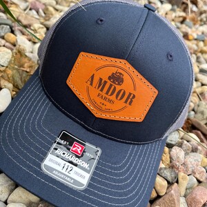 May include: A navy blue trucker hat with a gray mesh back. The front features a brown leather patch with the text "AMDOR FARMS" and a tractor graphic. The hat has a curved brim and a "Richardson" tag.