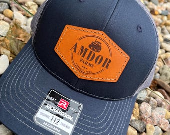Peut inclure: Casquette de camionneur bleu marine et grise avec un écusson en cuir portant l'inscription "AMDOR FARMS" et un dessin de tracteur. La casquette a une visière incurvée et une sangle réglable. L'étiquette indique "RICHARDSON ORIGINAL 112 TRUCKER".