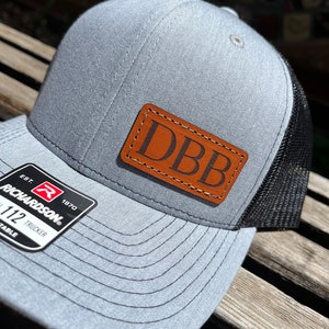 May include: A gray and black trucker hat with a brown leather patch that says "DBB" on it. The hat has a Richardson logo on the side.