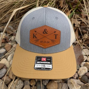 May include: A gray and tan trucker hat with a brown leather patch that says "K & T 07.13.24". The hat has a white mesh back and a brown leather patch with the Richardson logo and "Low-Pro 115 Trucker" text.
