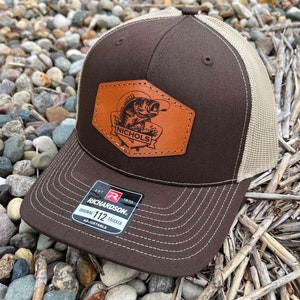May include: Brown and tan trucker hat with a brown leather patch featuring a fish and the text "Nichols". The hat has a Richardson 112 label and is adjustable.