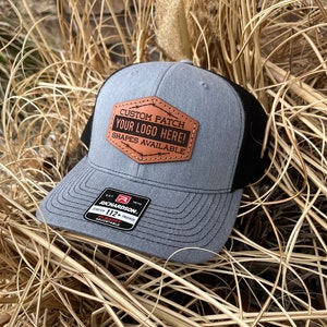 May include: A gray and black trucker hat with a brown leather patch that says "CUSTOM PATCH YOUR LOGO HERE! SHAPES AVAILABLE". The hat is made by Richardson and is a 112+ Trucker Flexfit.