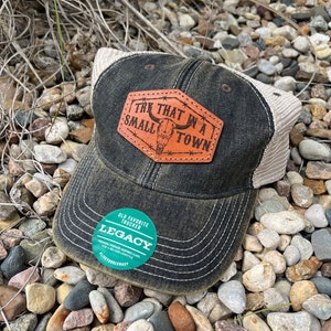 May include: A black and brown trucker hat with a brown leather patch that says "Try That In A Small Town" with a skull design. The hat has a mesh back and a green sticker that says "Old Favorite Trucker Legacy".