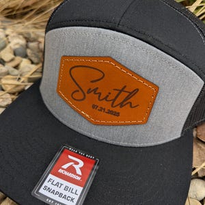May include: A black and gray baseball cap with a brown leather patch that says "Smith 07.21.2025". The cap has a mesh back and a flat bill. The cap has a red and white label that says "Richardson" and "Flat Bill Snapback Est 1970".