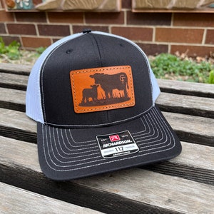 May include: Black and white trucker hat with a leather patch featuring a cow and calf silhouette. The hat has a curved brim and a white mesh back. The patch has the initials "GF" and the brand tag "Richardson 112 Trucker".