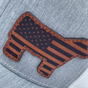 May include: A gray and white trucker hat with a brown leather patch in the shape of a cow with the American flag design.