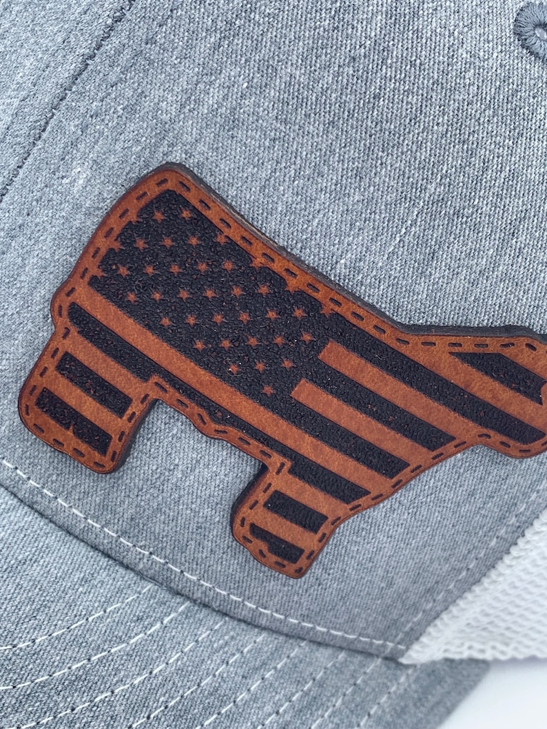 May include: A gray and white trucker hat with a brown leather patch in the shape of a cow with the American flag design.