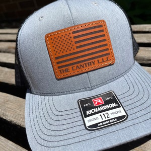 May include: Gray trucker hat with a black mesh back and a brown leather patch featuring an American flag design. The text "THE CANTRY L.L.C." is below the flag. The hat has a Richardson 112 Trucker tag.