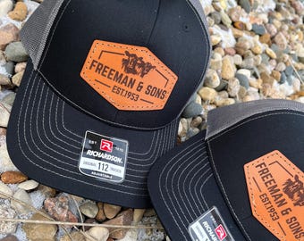 May include: Two black trucker hats with gray mesh backs. Each hat features a brown leather patch with the text "FREEMAN & SONS EST.1953" and a graphic of cattle. The hats have a curved brim and a tag that reads "RICHARDSON ORIGINAL 112 TRUCKER ADJUSTABLE."