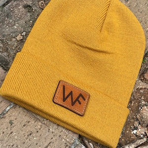 May include: Mustard yellow beanie hat with a folded brim. A brown leather patch with the letters "WF" is sewn on the front. The hat is made of a knitted material.