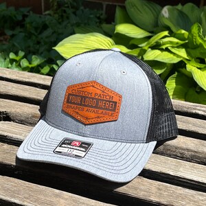 May include: Gray and black trucker hat with a brown leather patch. The patch reads "CUSTOM PATCH YOUR LOGO HERE! SHAPES AVAILABLE." The hat has a curved brim and a mesh back. The brand tag reads "RICHARDSON 112 ADJUSTABLE."