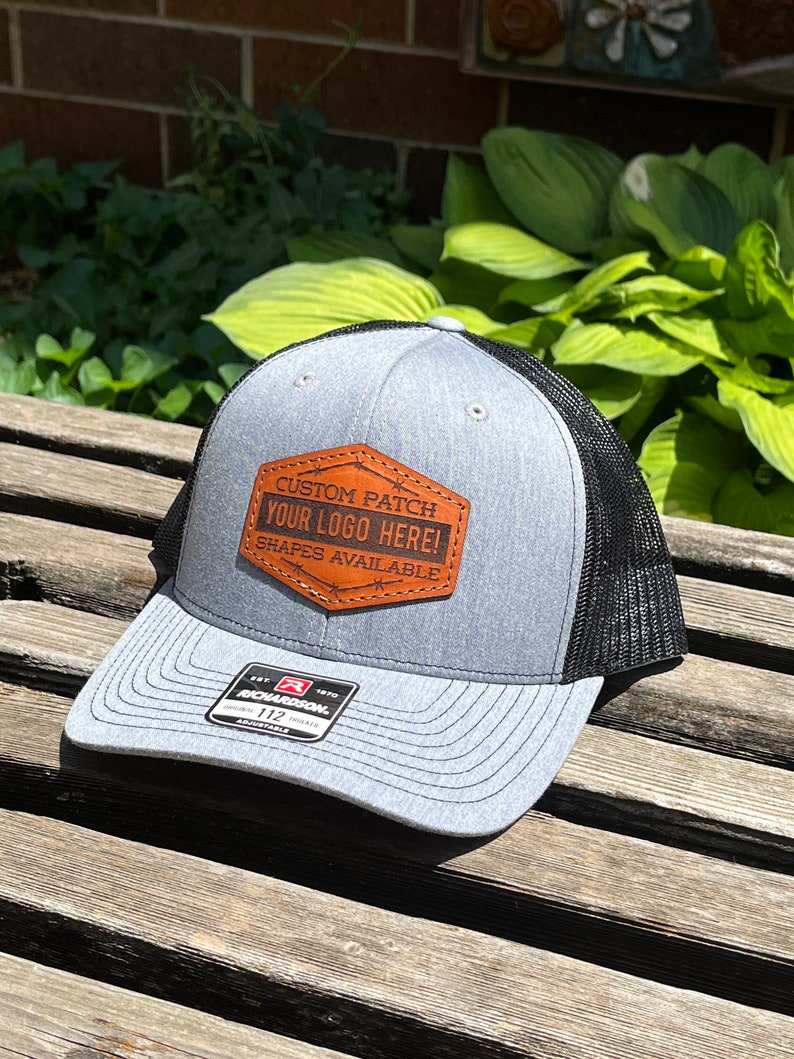 May include: Gray and black trucker hat with a brown leather patch. The patch reads "CUSTOM PATCH YOUR LOGO HERE! SHAPES AVAILABLE." The hat has a curved brim and a mesh back. The brand tag reads "RICHARDSON 112 ADJUSTABLE."
