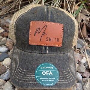 May include: A dark green and brown trucker hat with a brown leather patch that says "Mr. SMITH" in cursive. The hat has a green and white circular patch that says "LEGACY OFA VISOR BOARD MADE FROM RECYCLED CONTENT LIVE YOUR LEGACY".