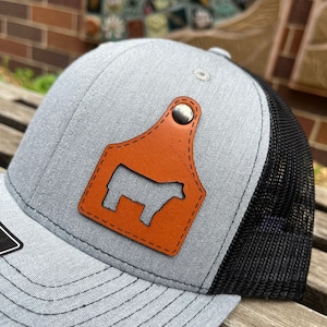 May include: A gray and black trucker hat with a brown leather patch featuring a silhouette of a cow. The patch has a silver rivet.