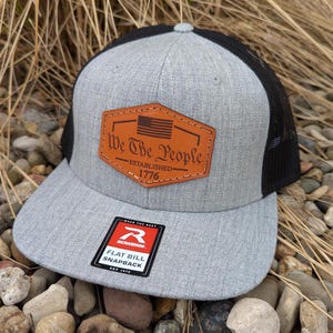May include: A gray and black trucker hat with a brown leather patch that says "We The People Established 1776" and a small American flag. The hat has a flat bill and a mesh back.