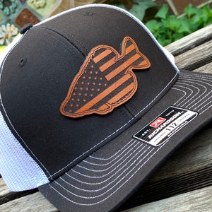 May include: A black and white baseball cap with a brown leather patch shaped like a fish with an American flag design. The patch is on the front of the cap. The cap is made by Richardson 112.