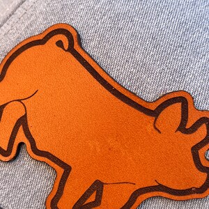 May include: A brown leather patch in the shape of a bear, with a dark brown outline, is affixed to a light blue denim fabric. The bear patch is the focal point, showcasing a simple, stylized design. The denim provides a textured background.