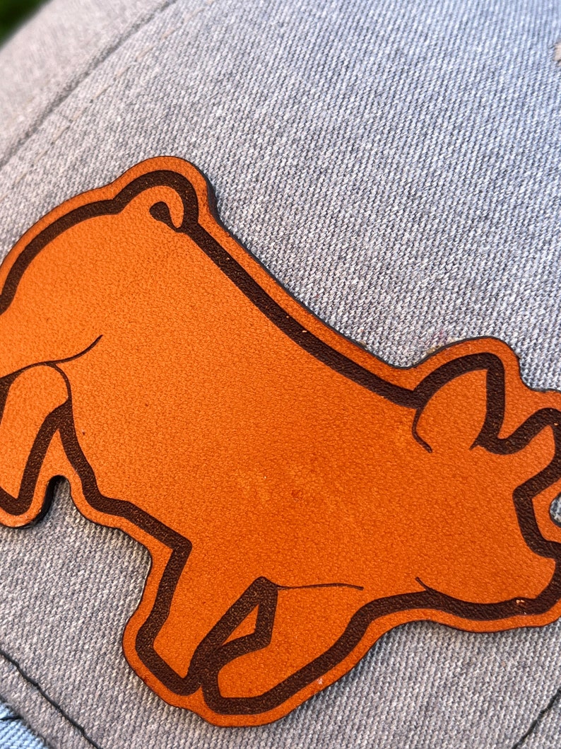 May include: A brown leather patch in the shape of a bear, with a dark brown outline, is affixed to a light blue denim fabric. The bear patch is the focal point, showcasing a simple, stylized design. The denim provides a textured background.