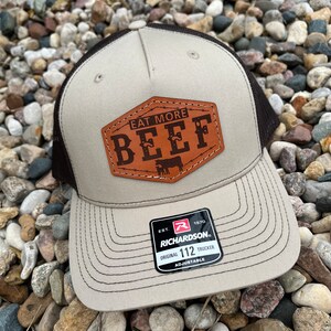May include: A brown and black trucker hat with a brown leather patch that says "Eat More Beef". The hat is adjustable and has the Richardson 112 logo on the back.