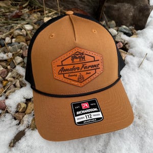 May include: A brown and black trucker hat with a leather patch that says "Amdor Farms" and a cow silhouette. The hat is adjustable and has the Richardson logo on the side.