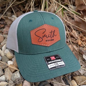 May include: A green and gray trucker hat with a brown leather patch that says "Smith 07.21.2025". The hat has a Richardson logo and the text "Low-Pro 115 Trucker MD/LG".
