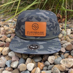 May include: A camouflage pattern baseball cap with a brown leather patch featuring the text "EVERBASE SOLUTIONS". The cap has a curved brim and a mesh back. The Osprey logo is on the brim.