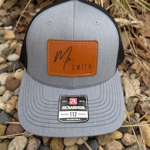 May include: Gray and black trucker hat with a leather patch that reads "Mr. Smith" in a script font. The hat has a curved brim and an adjustable strap. The Richardson brand tag is visible.