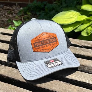 May include: A gray trucker hat with a black mesh back and a curved brim. The hat features a brown leather patch with the text "CUSTOM PATCH YOUR LOGO HERE! SHAPES AVAILABLE". The hat has a Richardson 112 tag.