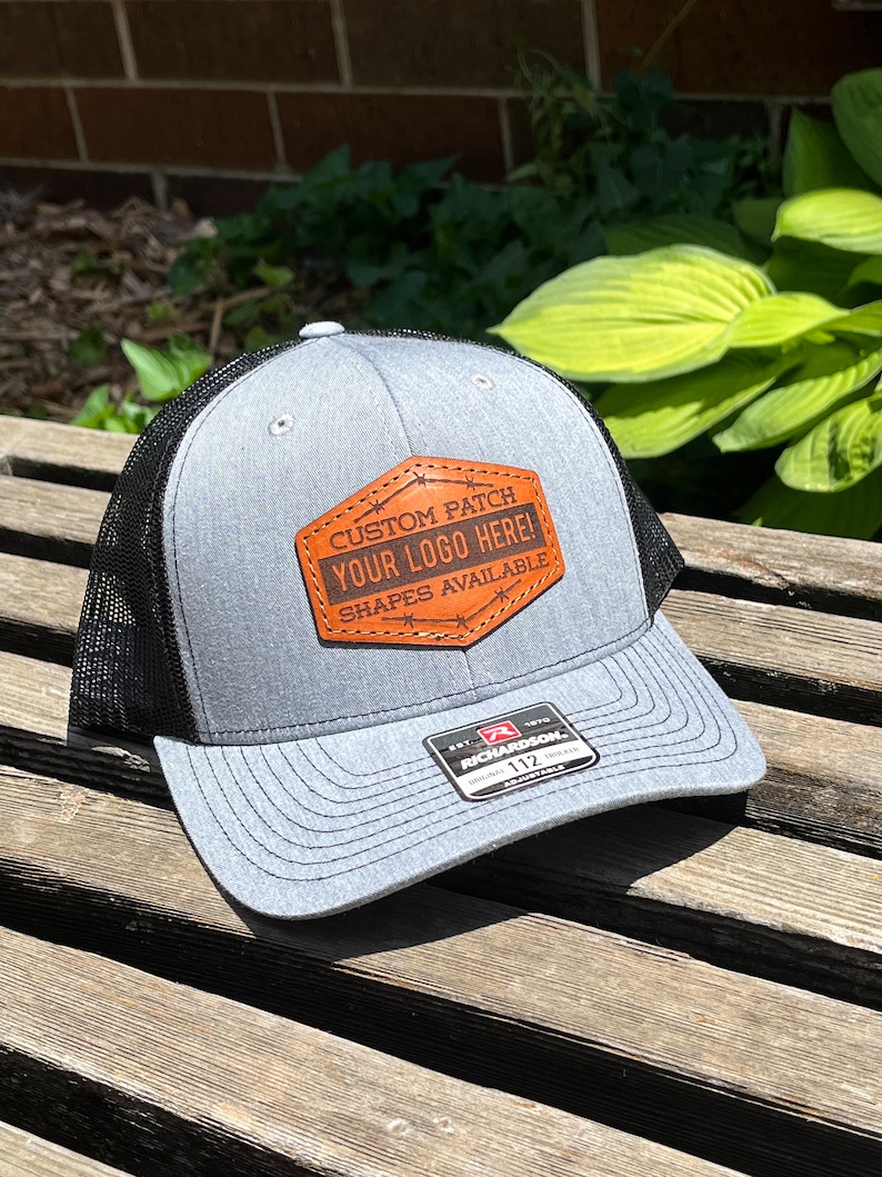May include: A gray trucker hat with a black mesh back and a curved brim. The hat features a brown leather patch with the text "CUSTOM PATCH YOUR LOGO HERE! SHAPES AVAILABLE". The hat has a Richardson 112 tag.