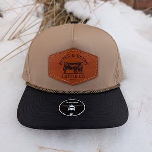 May include: A tan and black baseball cap with a leather patch featuring the text "ESTES & ESTES CATTLE CO." and an illustration of a cow and calf. The cap has a mesh back and a black brim.