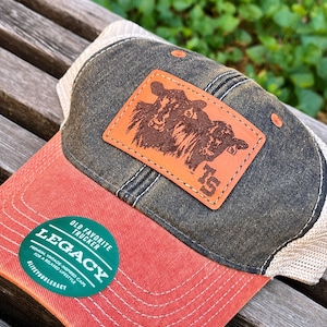 May include: A black and brown trucker hat with a red bill. The hat has a brown leather patch with a picture of two cows and the number 15. The hat also has a green patch that says "Old Favorite Trucker Legacy Premium Vintage-Inspired Caps for a Relaxed Lifestyle Live Your Legacy".