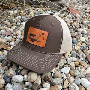 May include: A brown and tan baseball cap with a brown leather patch featuring a silhouette of a dog and ducks. The patch is stitched onto the front of the cap.