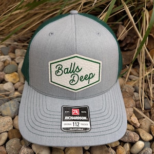 May include: A gray and green trucker hat with a white patch that says "Balls Deep" in a cursive font. The hat is made by Richardson 112 and is adjustable.