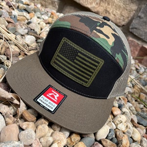 May include: A green, black, and gray camouflage baseball cap with a black mesh back. The cap has a flat bill and a small American flag patch on the front. The cap also has a red and white label that says "Richardson Flat Bill Snapback".
