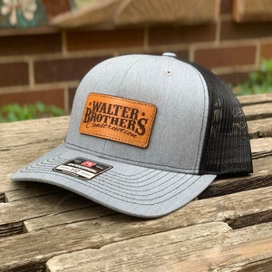 May include: A gray and black trucker hat with the text "WALTER BROTHERS Construction" on a brown leather patch. The hat has a mesh back and a curved brim. The hat is sitting on a wooden surface.