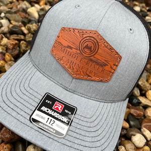 May include: A gray and black trucker hat with a brown leather patch that says "Middle River Carpentry". The hat has a black mesh back and an adjustable strap with a black buckle. The hat also has a red and white label that says "Richardson 112 Original Trucker Adjustable".
