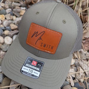 May include: A green and brown trucker hat with a brown leather patch that says "Mr. Smith". The hat has a mesh back and a Richardson logo on the side.