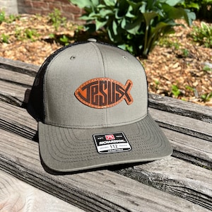 May include: A green and black trucker hat with a brown leather fish patch that says "JESUS" on it. The hat is adjustable and has a Richardson 112 label.