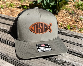 May include: Olive green trucker hat with a black mesh back. The hat features a brown leather patch in the shape of a fish with the word "JESUS" stitched on it. The hat is adjustable and has a Richardson brand tag.