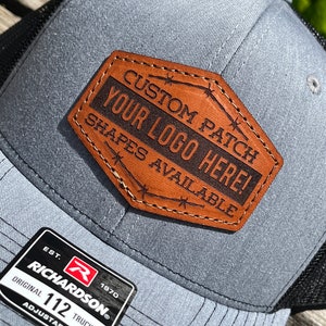 May include: Gray trucker hat with a black mesh back and a brown leather patch. The patch reads "CUSTOM PATCH YOUR LOGO HERE! SHAPES AVAILABLE." The hat also has a Richardson 112 Trucker tag.